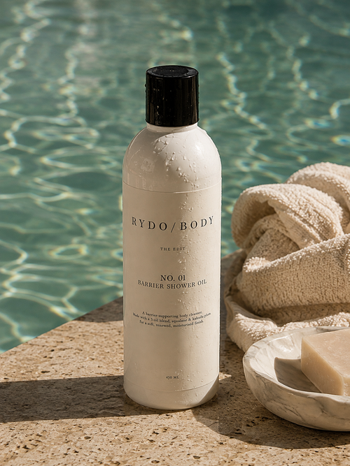 A bottle of Rydo Body The Rich No. 01 Barrier Shower Oil stands on a stone poolside surface with a folded beige towel and a small dish of soap in the background, with rippling pool water behind it.